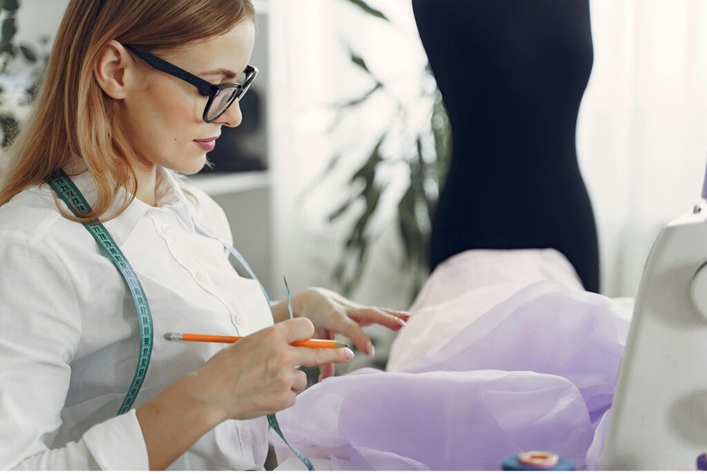 Woman measuring fabric and preparing alterations with a sewing machine and dress form in a well-lit sewing studio.